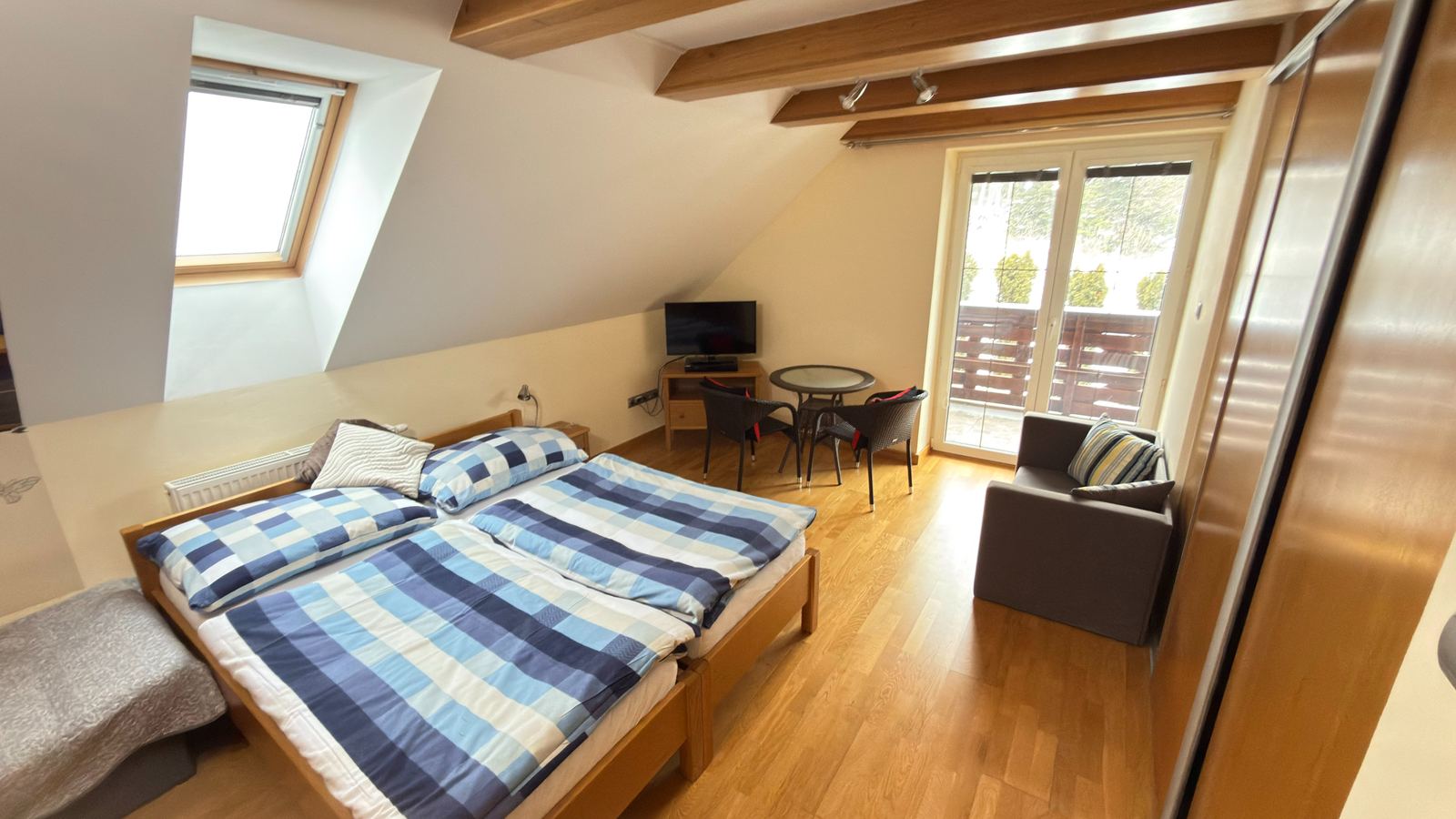 Svetlá podkrovná izba s manželskou posteľou, drevenými trámami, stolíkom a dverami na balkón